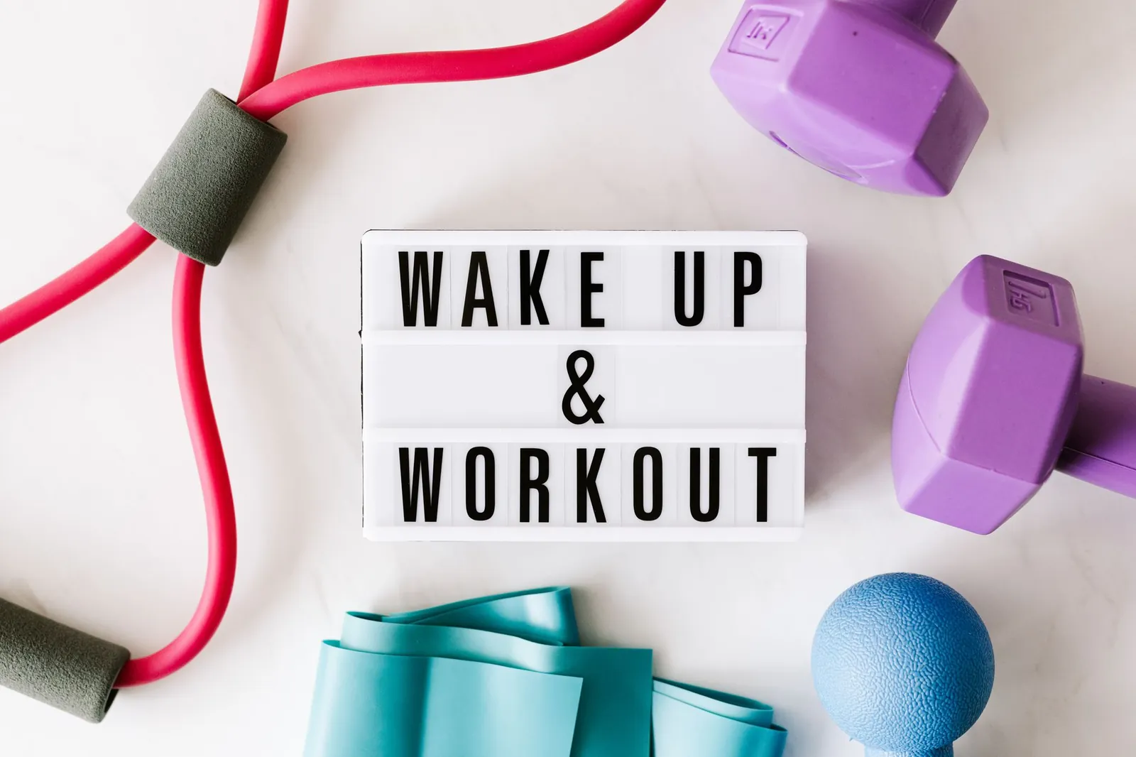 Five color-coded resistance bands coiled on a gym mat next to a pair of adjustable dumbbells on a compact rack, showing the size and storage contrast
